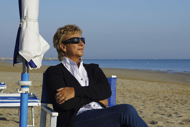 Frau mit Sonnenbrille sitzt in Stuhl am Strand und erholt sich.