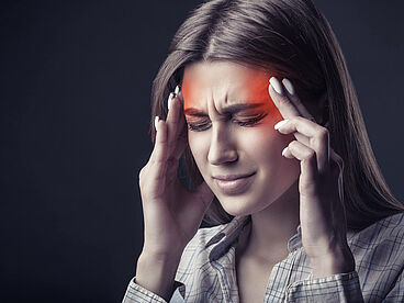 Eine Frau ist von Kopfschmerzen geplagt und fasst sich an die Schläfen. Der schmerzende Bereich ist rot eingefärbt.